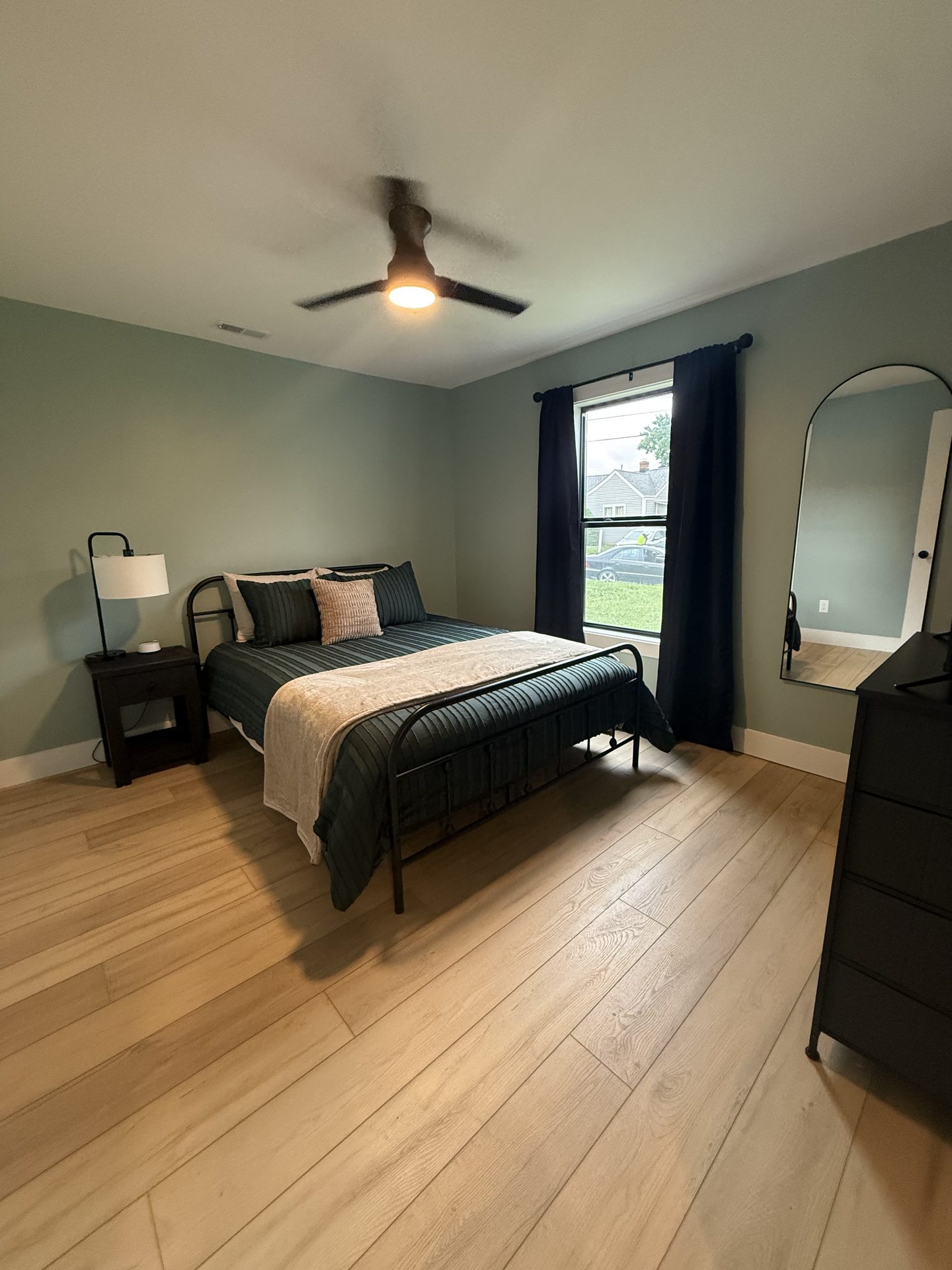 Bedroom painted soft green with a queen bed, blackout curtains, a mirror and a dresser