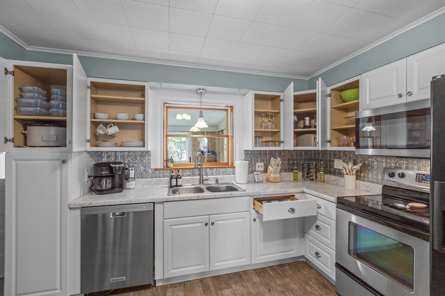 White kitchen with open cabinets showing dishes and glassware, a double sink under the window and a stainless range and dishwasher