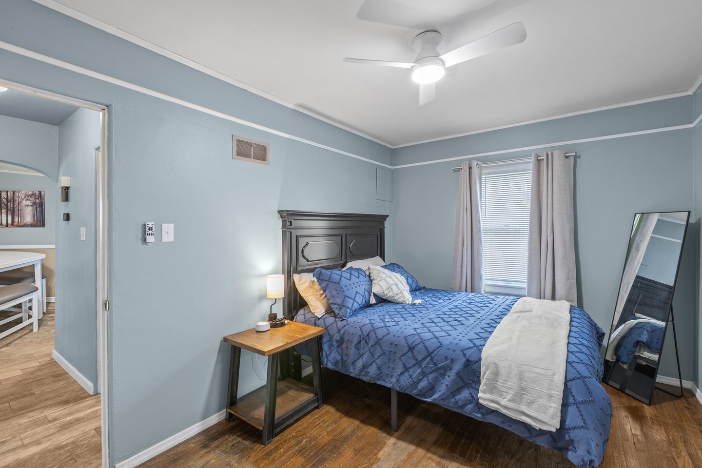 Bedroom with a queen bed in blue bedding, a dark headboard, a floor mirror and a tall window