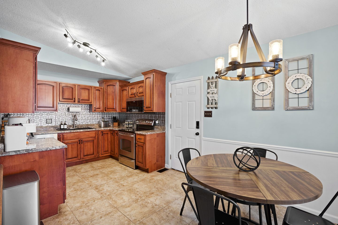 Round wooden dining table with metal chairs under a wagon-wheel chandelier, beside the kitchen