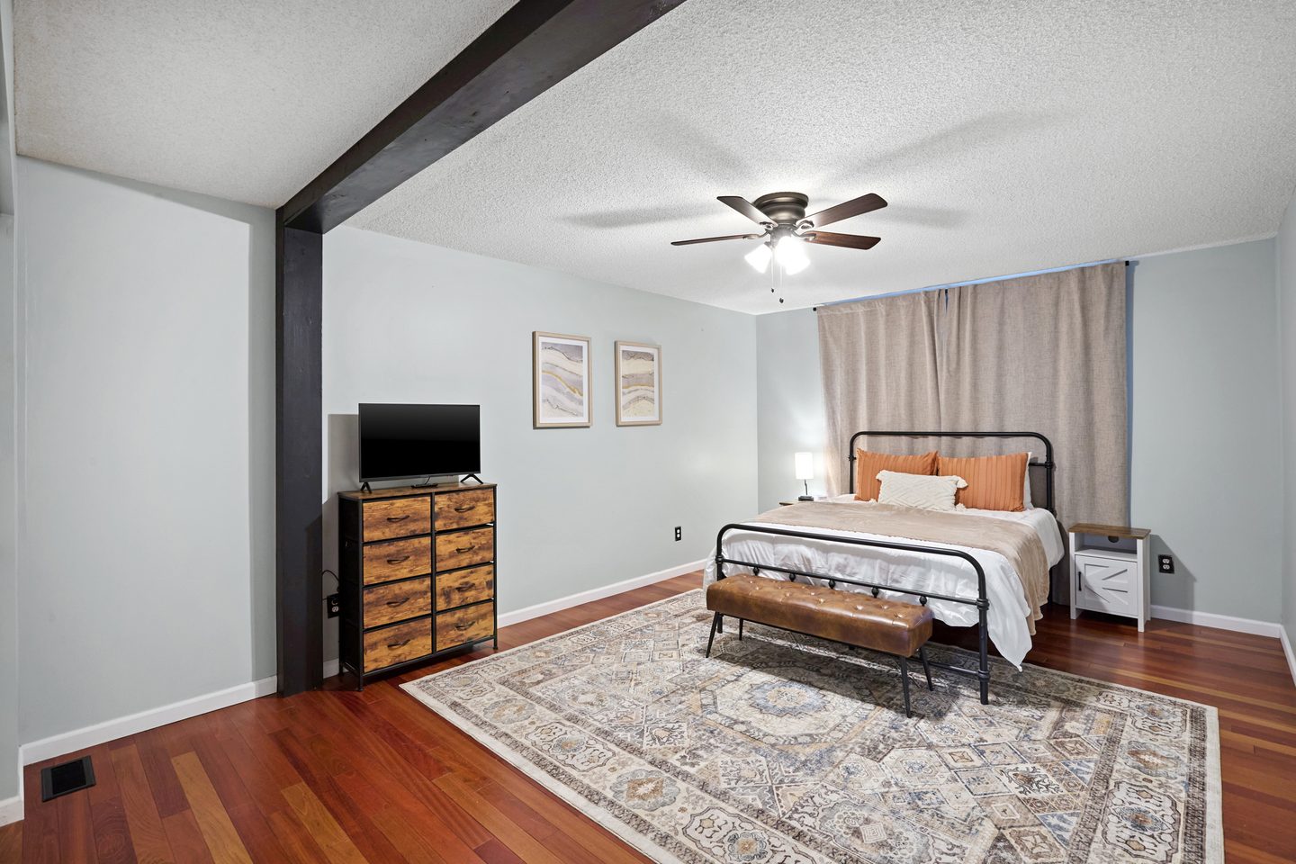 Bedroom with a queen bed on a black metal frame, a leather bench at its foot, a large rug and a dresser with a television