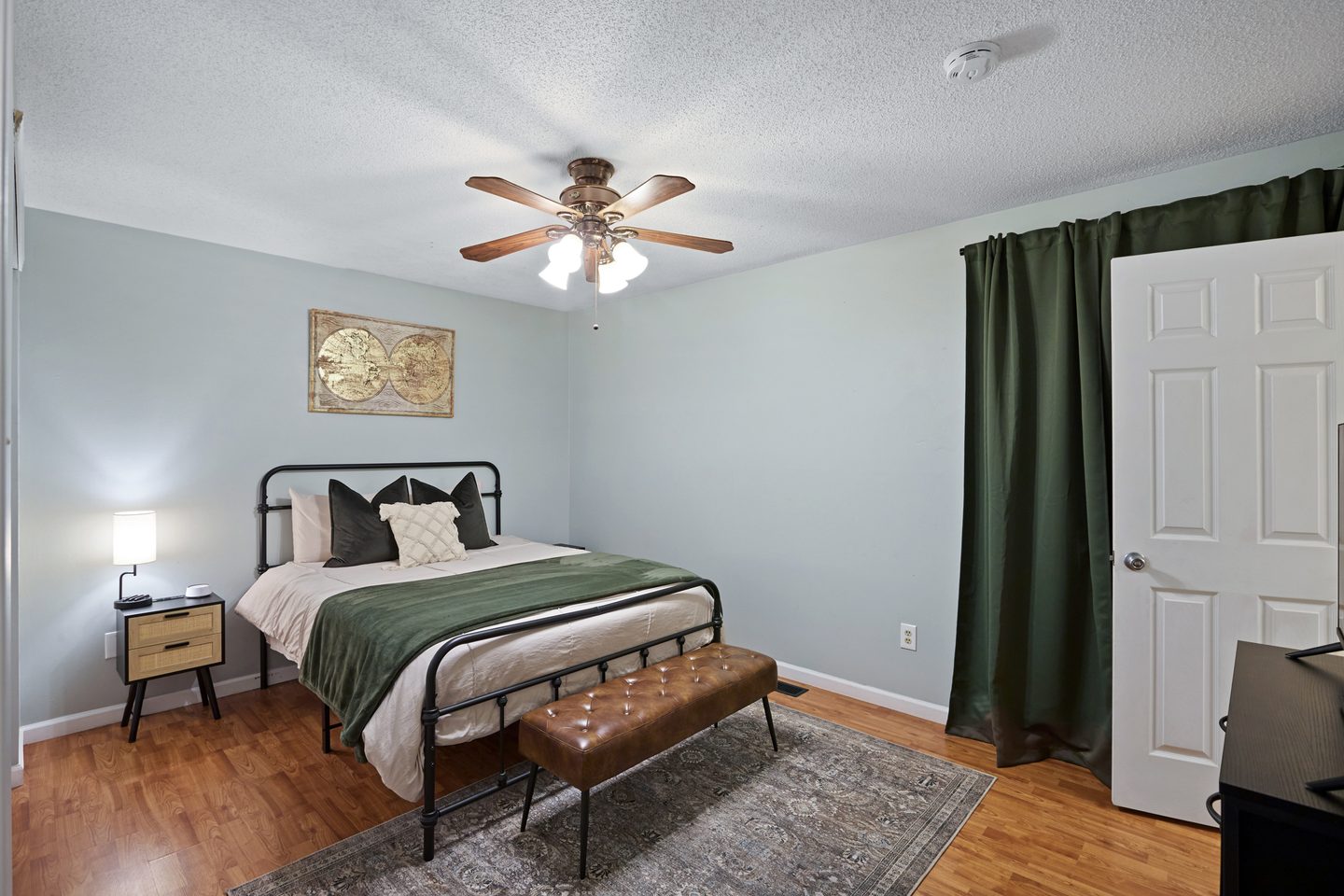 Bedroom with a queen bed in green bedding, a leather bench, a framed map and a ceiling fan