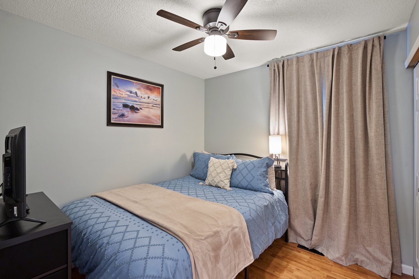 Bedroom with a queen bed in blue bedding, tall curtains, a ceiling fan and a television on a dresser