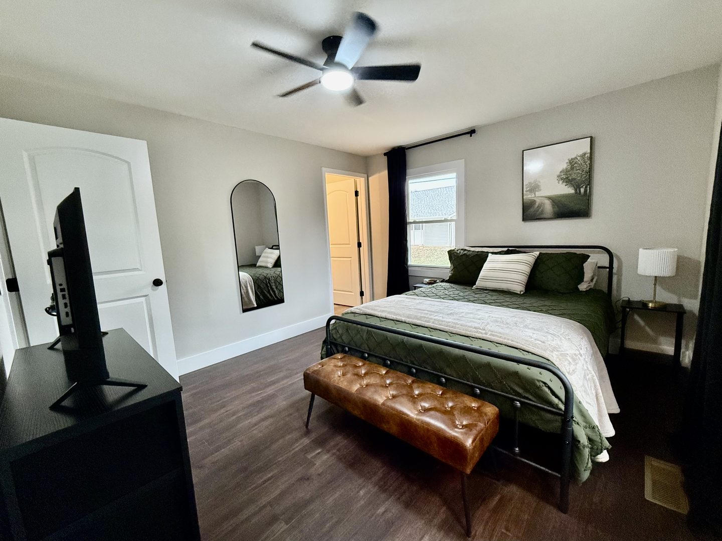 Bedroom with a queen bed in green bedding, a leather bench, an arched mirror and a television