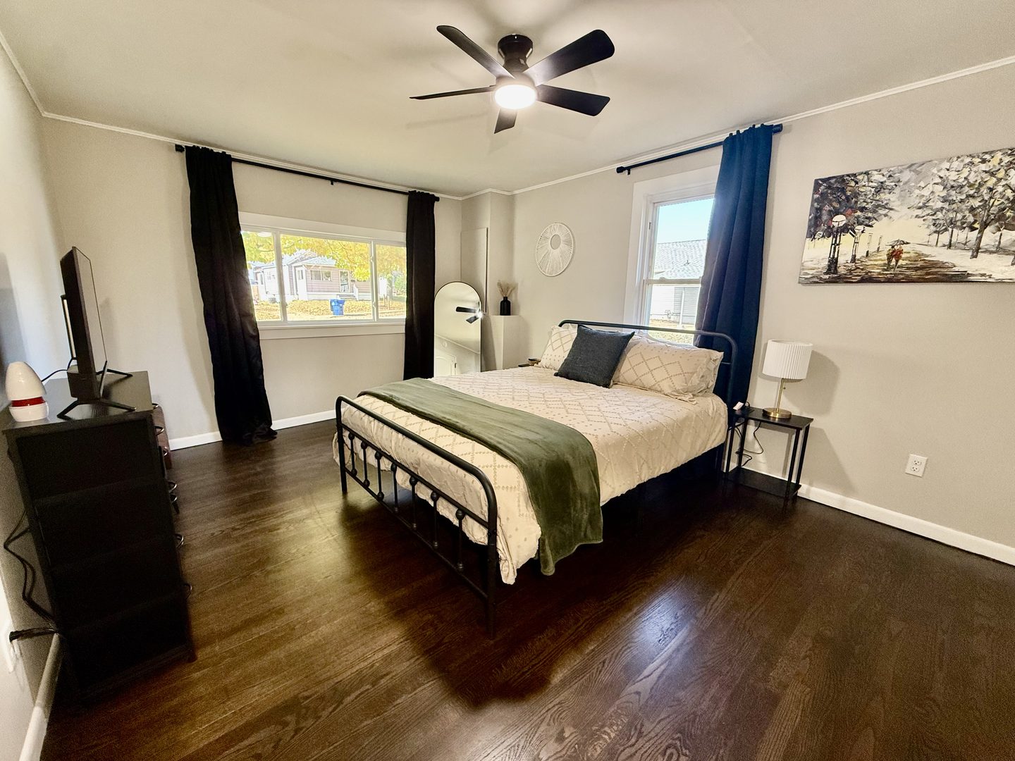 Bedroom with a queen bed on a black metal frame, two windows with dark curtains and a ceiling fan