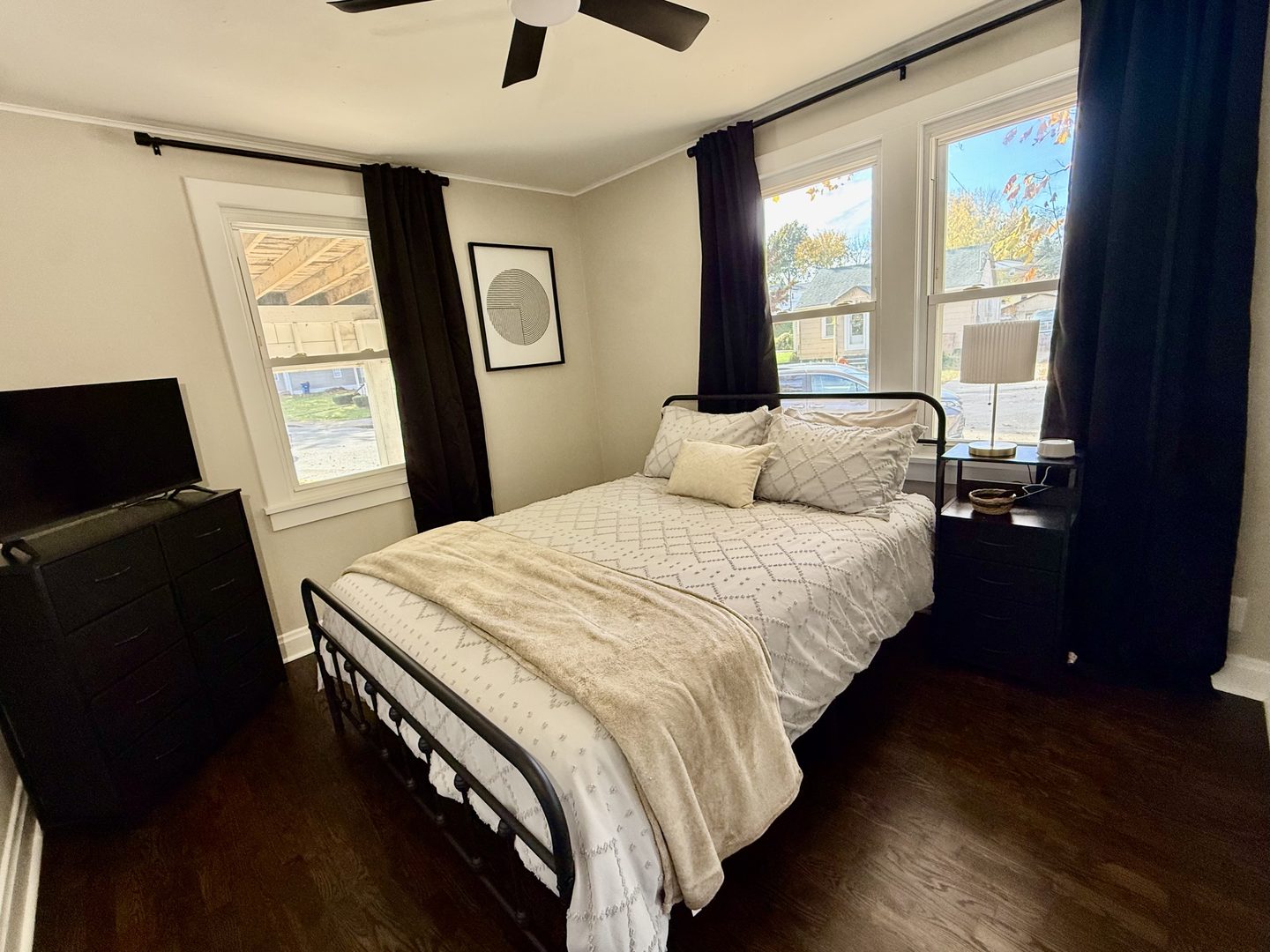 Bedroom with a queen bed in cream bedding, black curtains and a television on a dresser